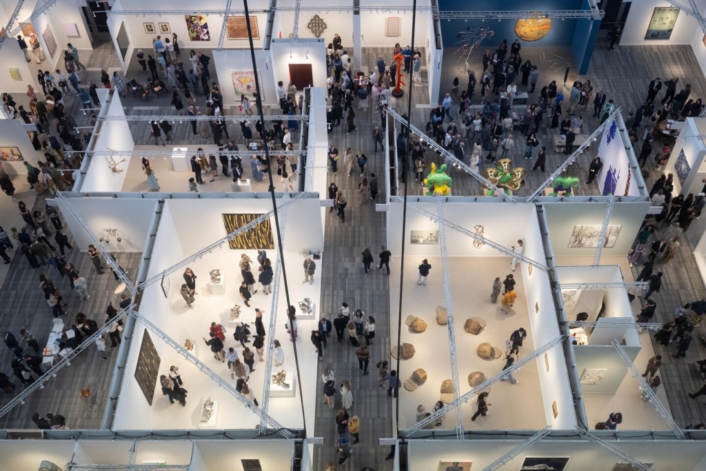 , An overhead, high-angle view of a large, busy art fair inside a modern hall. Multiple white-walled booths display various paintings and sculptures. Many people are walking through the aisles and looking at the artwork. Frieze New York