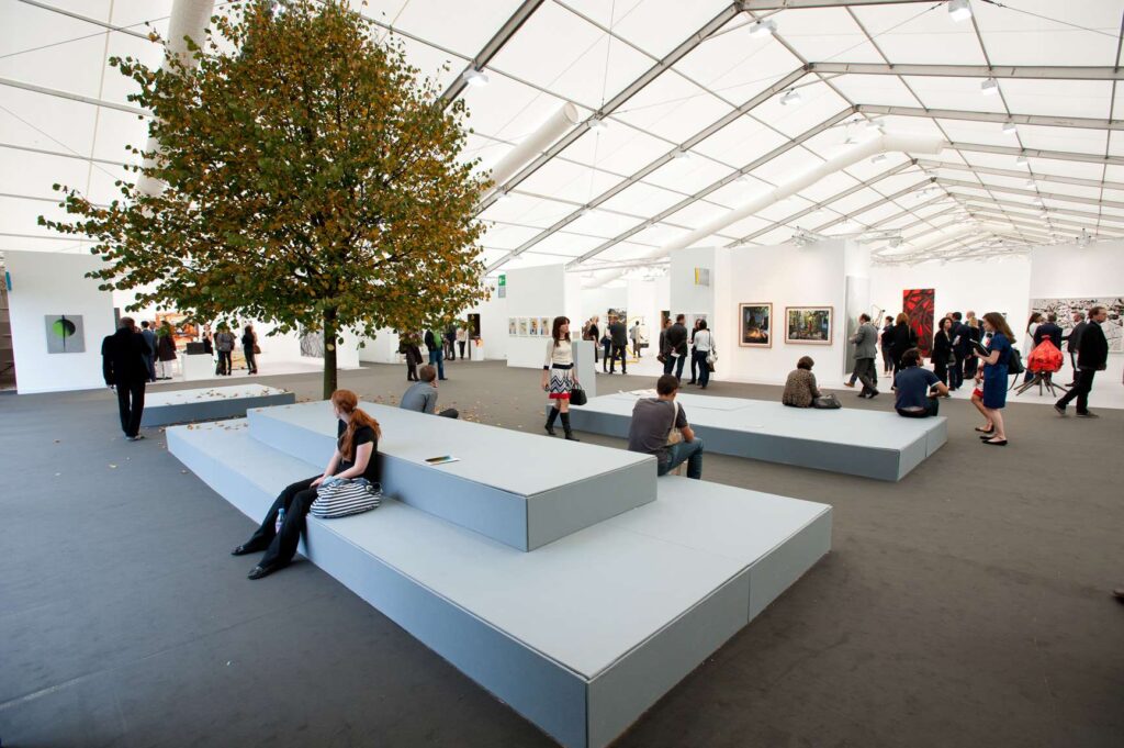 Frieze New York, The interior of a very large, white tent used for an art fair. There is a real tree growing in the middle of the room next to grey seating blocks. People are scattered throughout the space, visiting different art stalls in the distance.