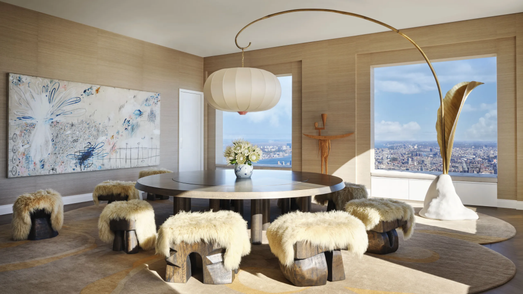 A luxury dining area featuring unique, log-style stools covered in light sheepskin, grouped around a dark, circular wooden table. A large, stylized floor lamp with a golden stem and massive leaf-shaped shade curves over a corner. Large windows offer panoramic city and water views. Original art by interior designers