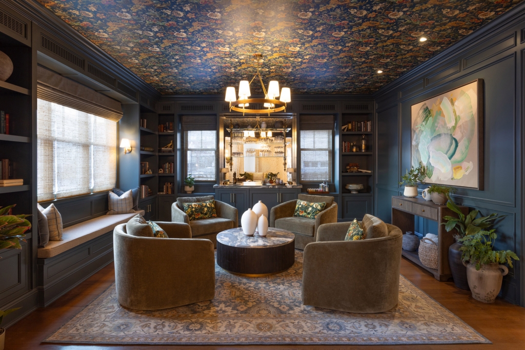 Intimate and sophisticated lounge with walls painted in a deep dark-blue shade and a ceiling covered with a richly detailed floral wallpaper, furnished with four velvet armchairs arranged around a marble coffee table and illuminated by a golden circular chandelier and classic sconces, interior designers.