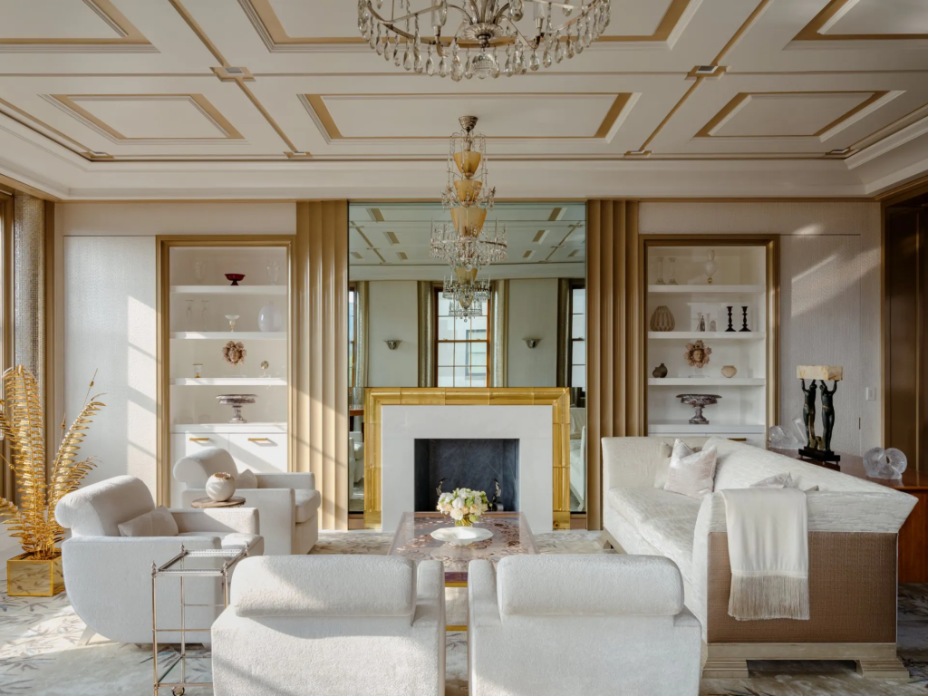 An elegant, bright white living room with gold trim detailing on the ceiling, featuring a white sofa, white armchairs, a gold-framed fireplace with a mirror above it, and two crystal chandeliers interior designers