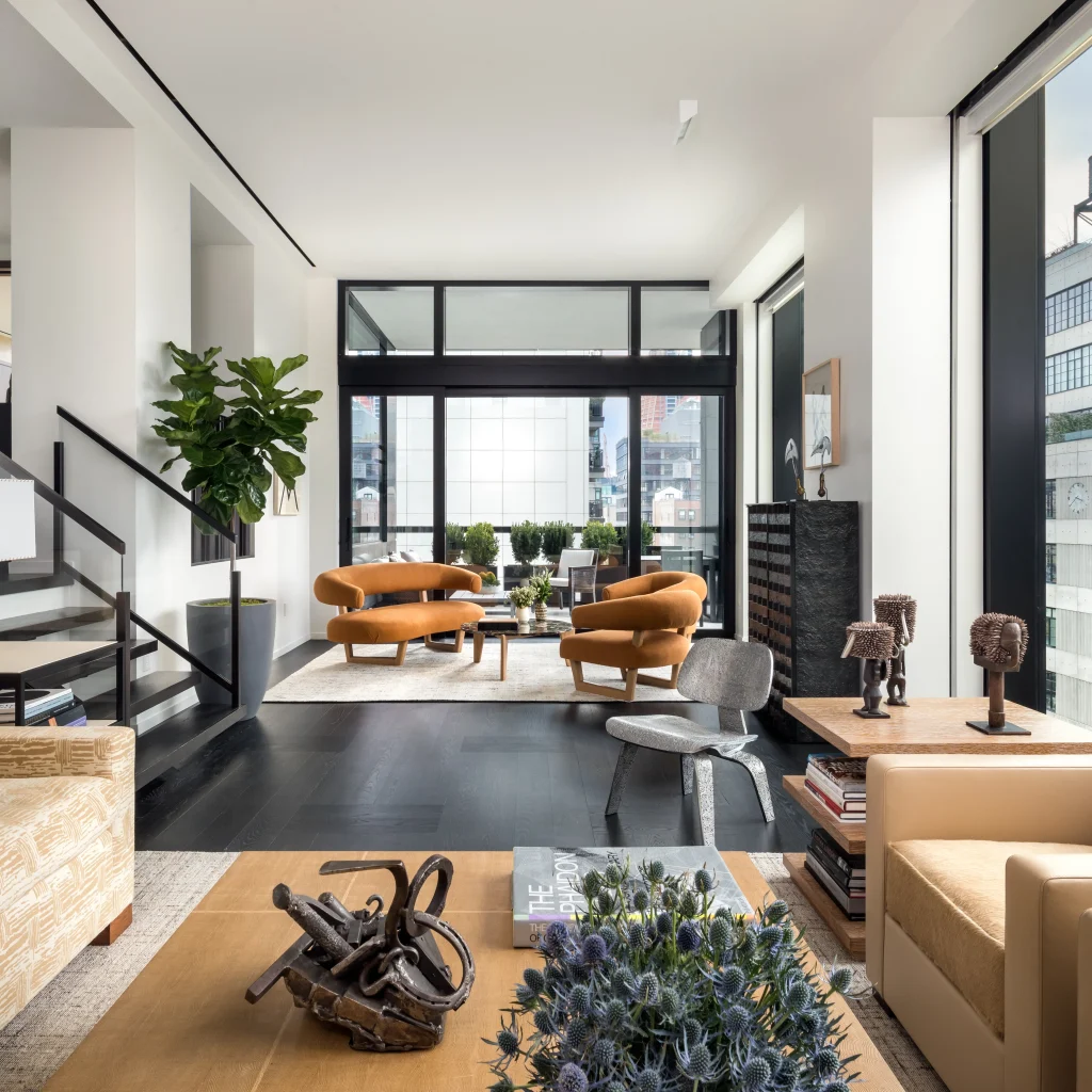A contemporary two-story loft apartment living space with a second-floor balcony over black metal stairs. Curved, burnt orange sofas define the main seating area on a textured rug. Floor-to-ceiling glass doors open to an outdoor terrace with potted plants and city views. Various artistic objects, sculptures, and furniture by interior designers