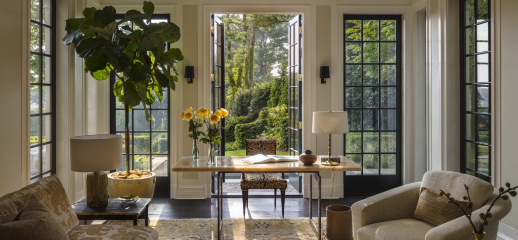 A classic, well-lit study with black steel-framed French doors and windows looking out onto a lush garden. A polished wood desk is paired with a leopard-print chair. Two different table lamps sit on side surfaces. A large Fiddle Leaf Fig tree in a yellow pot stands in one corner. Room styled by interior designers