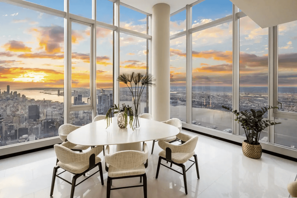 Modern high-rise dining room with a white triangular table and cream-colored design chairs, positioned in front of immense glass walls that reveal a stunning aerial view of a coastal metropolis during sunset, interior designers.