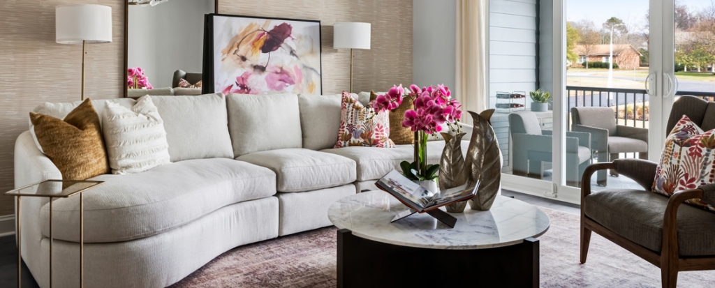Cozy living room with an off-white curved sofa decorated with gold and floral patterned pillows, a round marble coffee table with a vase of pink orchids, and sliding glass doors opening onto an outdoor balcony, interior designers.