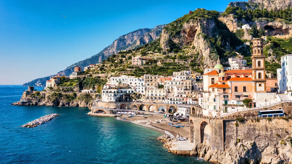A sunny view of a colorful coastal town built onto a steep cliffside next to a sandy beach, Coastal Roads.