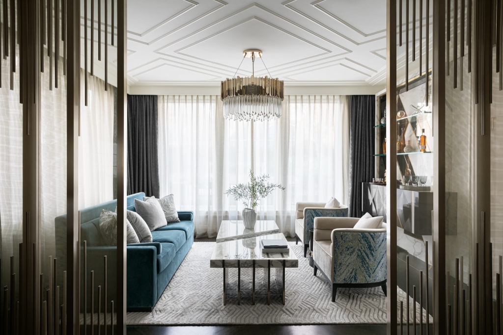 A view through a decorative screen into a stylish living room with a geometric ceiling pattern, featuring a long teal velvet sofa, two patterned armchairs, a marble coffee table, and a crystal chandelier interior designers