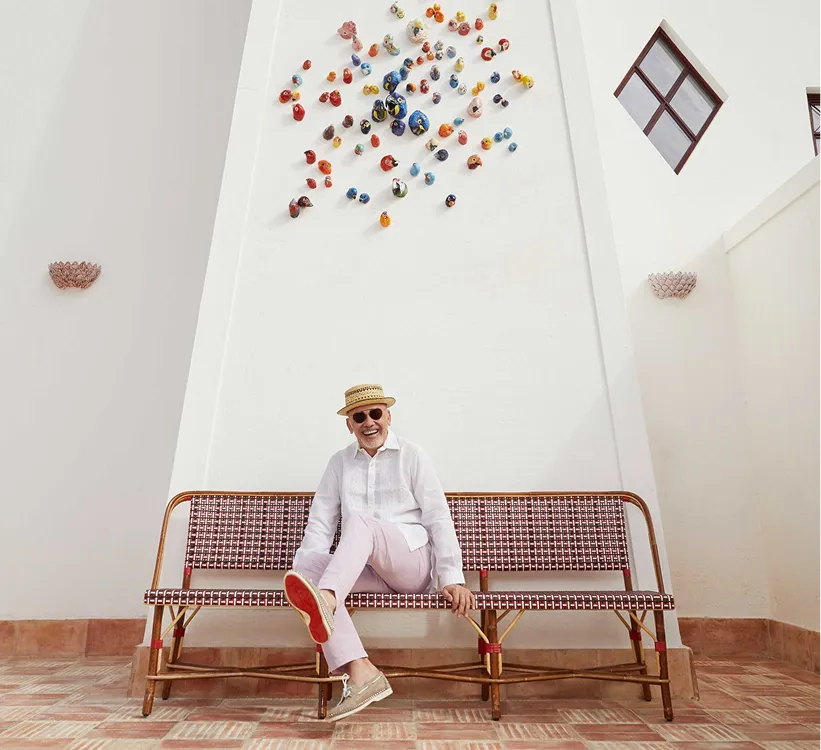 A smiling man in a white shirt, pink trousers, sunglasses, and a straw hat sits cross-legged on a patterned bench against a tall white wall decorated with small, colorful ceramic bird figures. Vermelho Hotel