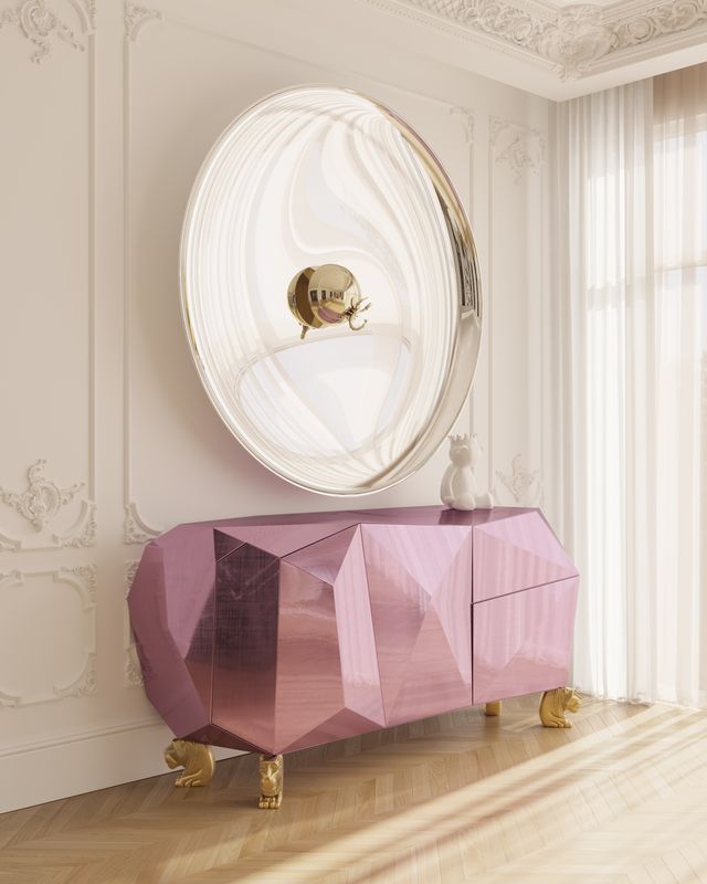 A bright room corner showcasing a futuristic, faceted pink credenza with gold lion-paw feet. Hanging above the sideboard is a large, circular convex mirror reflecting the room, set against a classic white paneled wall near a window with sheer curtains. luxury bedroom
