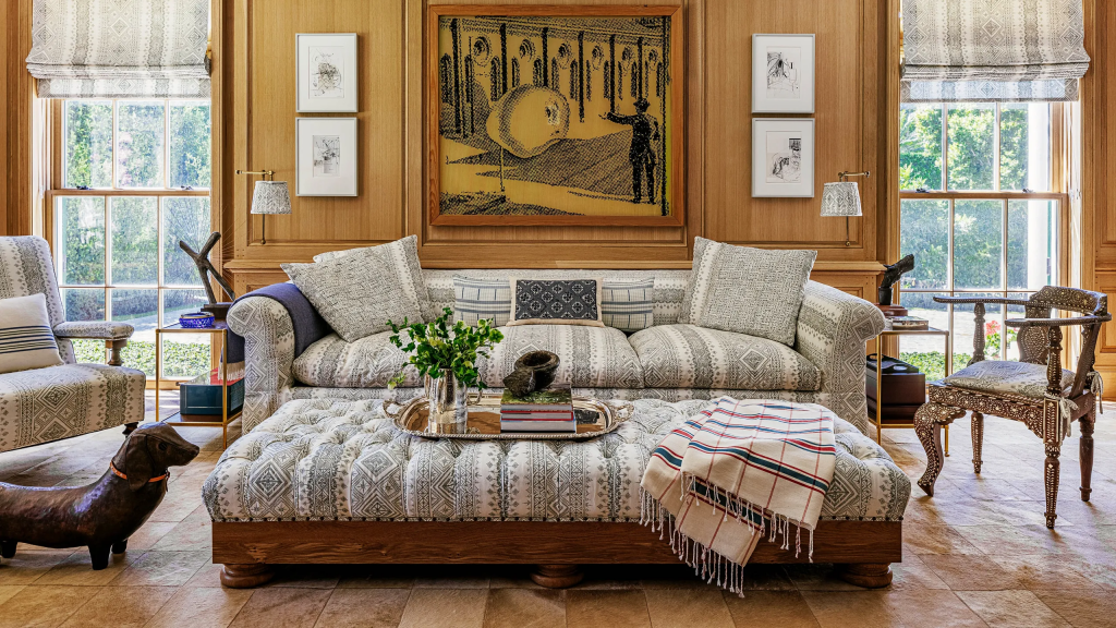 A cozy living room with light wood paneled walls, featuring a large tufted ottoman and a matching sofa upholstered in a light grey patterned fabric, accented by a large abstract painting and framed artwork on the wall interior designers