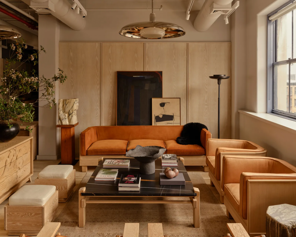A warm, mid-century modern sitting area with light wood walls, featuring an orange velvet sofa, two matching leather armchairs, a black marble coffee table with art books, and minimalist sculptures interior designers