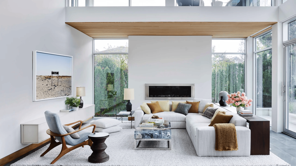 Contemporary and bright living room with white walls, a large light-colored modular L-shaped sofa with throws and pillows in earthy tones, a vintage-style wooden armchair, and wide glass windows integrating the indoor space with the view of a lush green garden in the background, interior designers.