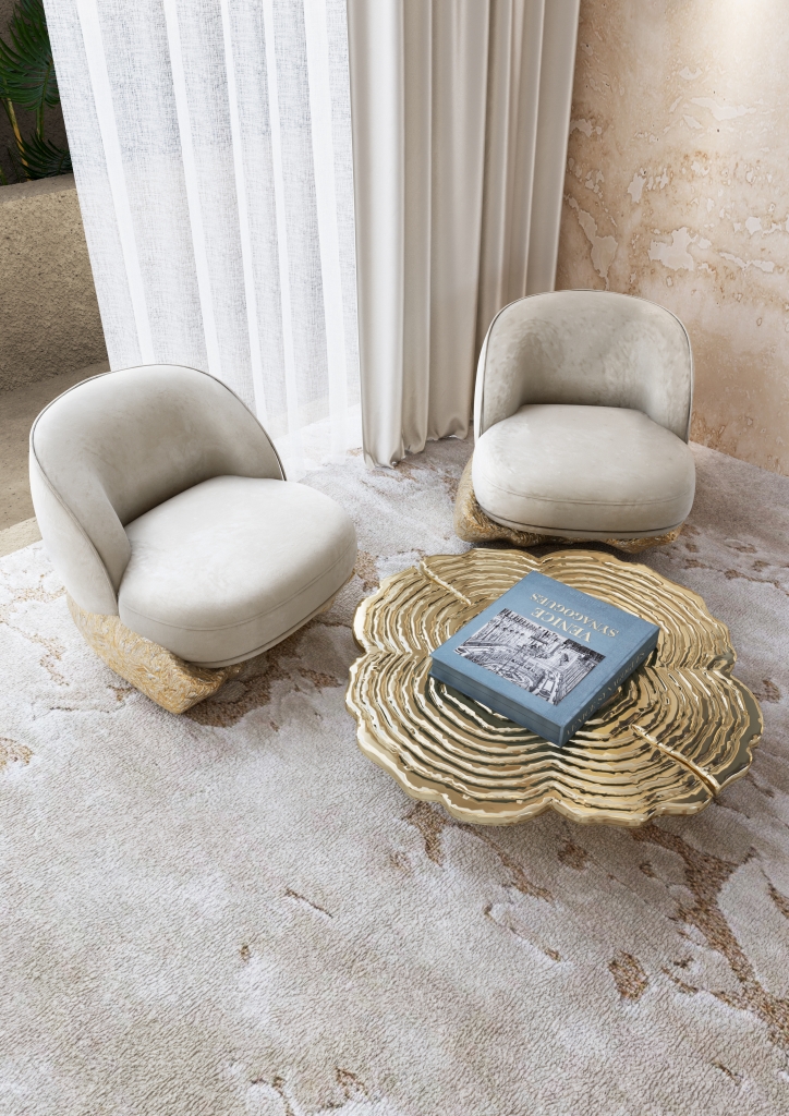 Aerial view of two ivory velvet low armchairs with sculpted gold bases flanking a large gold rippled coffee table on a textured cream rug, beside sheer white floor-to-ceiling curtains living rooms