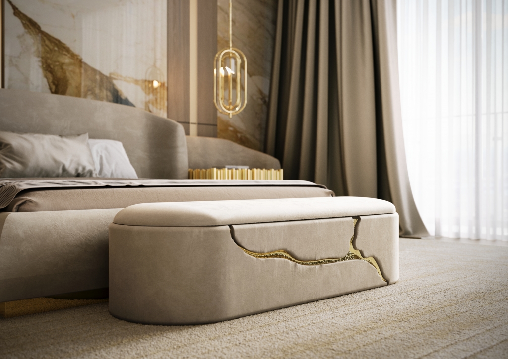 A close-up shot of a contemporary bedroom focusing on an elegant cream-colored upholstered bench. The bench is supported by organic, sculptural bases finished in polished gold, mimicking the shape of natural rocks, set against a soft beige carpet and bed frame. luxury bedroom