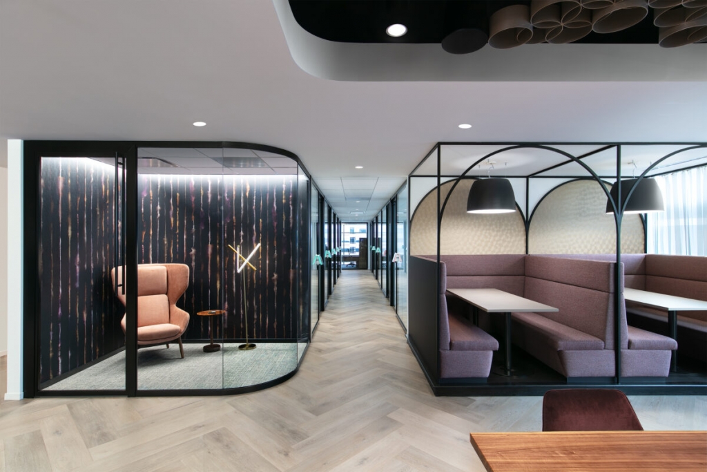 A modern office hallway with herringbone wood flooring, featuring a glass-enclosed meeting room on the left with a pink armchair, and private dining or meeting booths with purple seating on the right under large black pendant lights interior designers