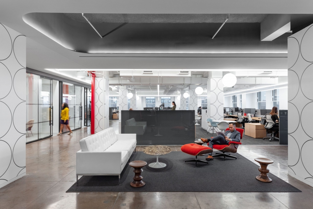 An open-plan corporate office lounge area with polished concrete floors, featuring a white leather sofa, a marble coffee table, and a person sitting on a red lounge chair with an ottoman reading, surrounded by columns with geometric patterns interior designers