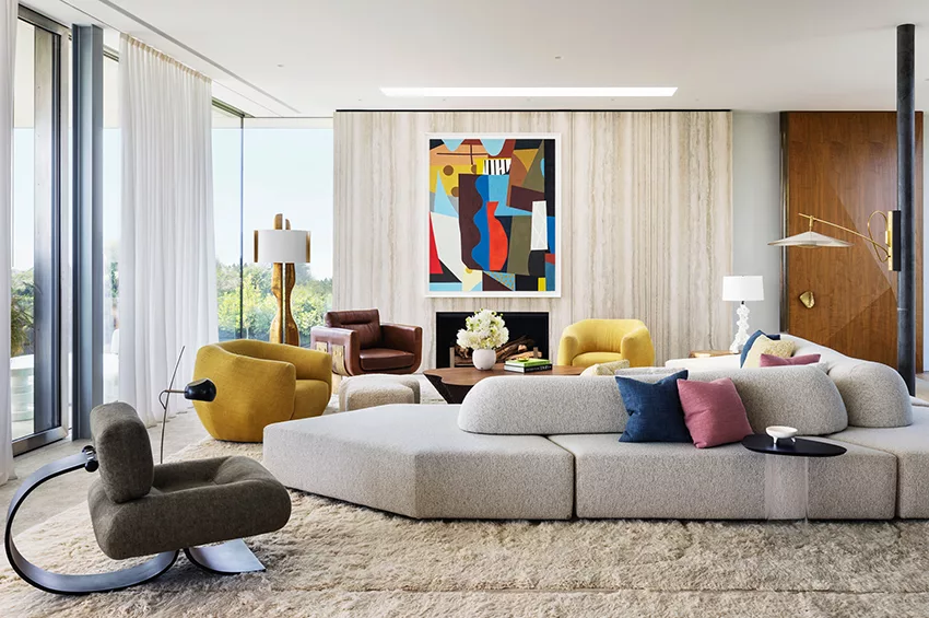 A bright, contemporary living space with an expansive neutral sectional sofa, colorful throw pillows, bold abstract artwork above a fireplace, and a textured rug, decorated by interior designers