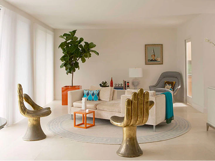 A bright, modern living room showcasing two large, sculpted, brass hand-shaped chairs as a focal point. A white sofa sits in the center on a patterned rug. A tall Fiddle Leaf Fig tree in an orange square planter stands beside a window. Art and diverse decor, including small, orange side tables, complete the space designed by interior designers