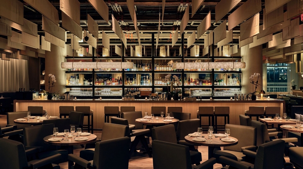 An upscale restaurant dining room with dark tables and chairs, a long illuminated bar backed by shelves of bottles, and decorative hanging fabric banners overhead, crafted by interior designers