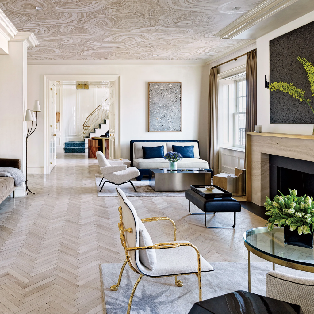 A bright, luxurious living room featuring herringbone wood floors, a textured marble pattern ceiling, elegant seating with a gold-framed armchair, and a classic fireplace mantel, curated by interior designers