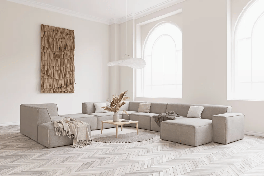 Minimalist and bright environment with herringbone pattern flooring, featuring a large light-grey modular U-shaped sofa arranged around a low wooden coffee table with a dried floral arrangement, under large arched windows and a fluid contemporary pendant light, interior designers.