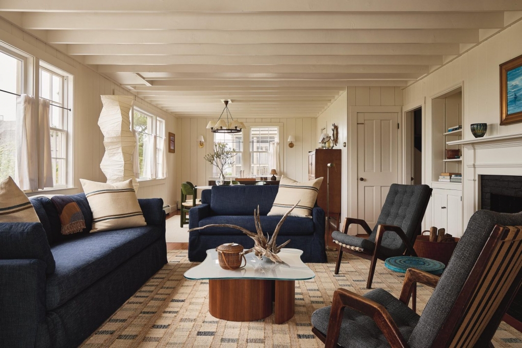 A bright, rustic living room with exposed white ceiling beams, featuring two deep blue sofas with striped pillows, a glass-top wooden coffee table, grey mid-century modern armchairs, and large windows interior designers