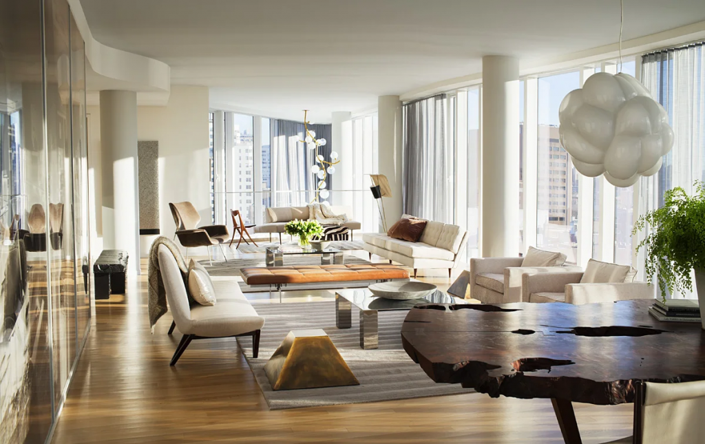 A spacious, modern open-concept apartment with floor-to-ceiling windows showing city views, filled with a large wooden dining table, contemporary sofas, and a cloud-like pendant light, styled by interior designers