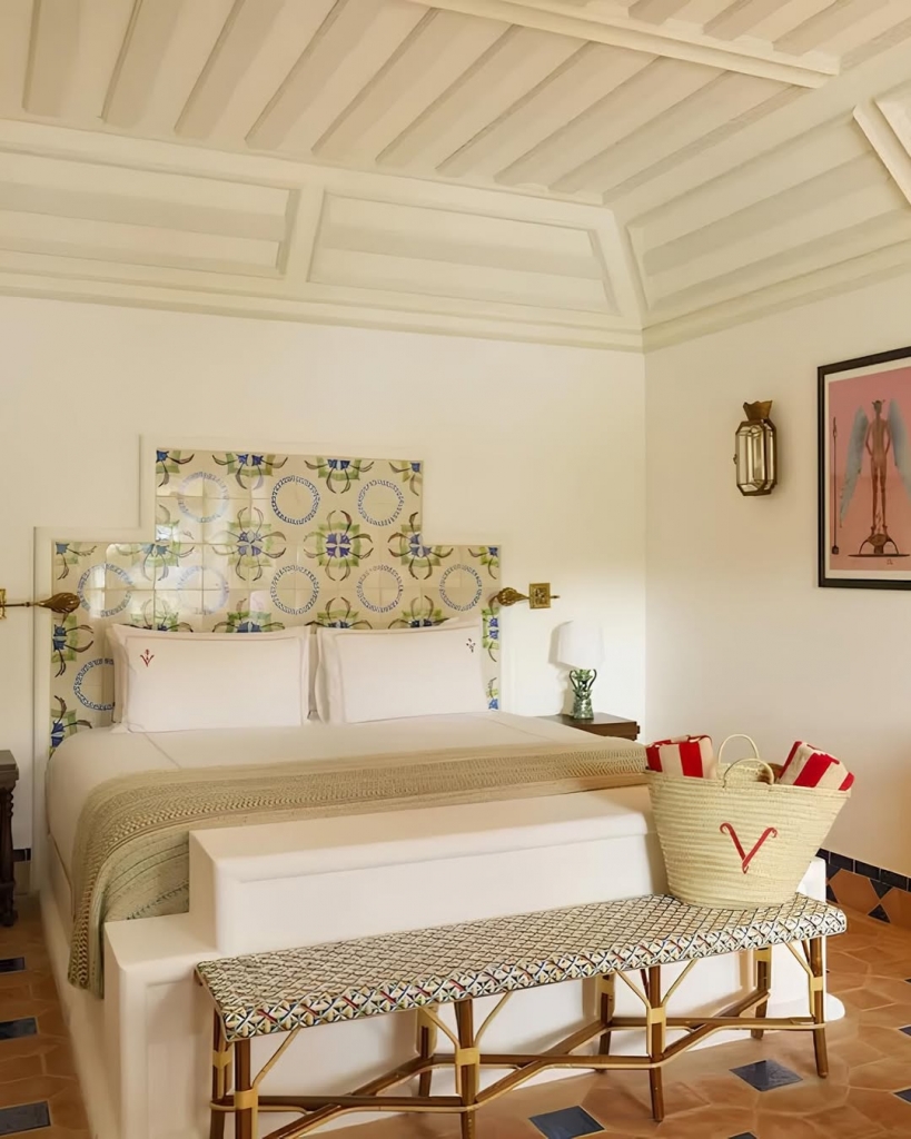 A bright bedroom featuring a white bed with a tiled headboard, a woven bench at the foot of the bed holding a beach bag, and terracotta floor tiles. Vermelho Hotel