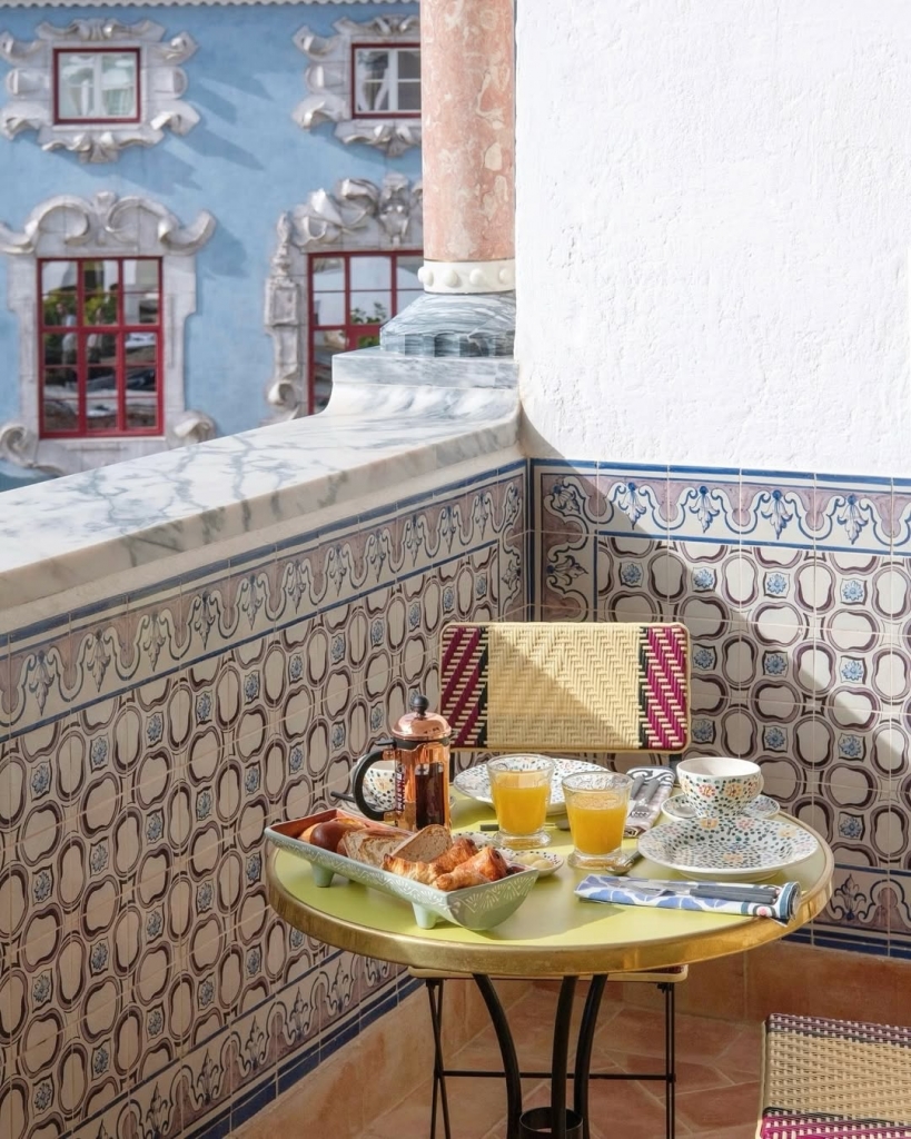 A sunny breakfast setup for two on a balcony table with orange juice, coffee, and pastries, set against a wall of traditional blue and white Portuguese tiles. Vermelho Hotel