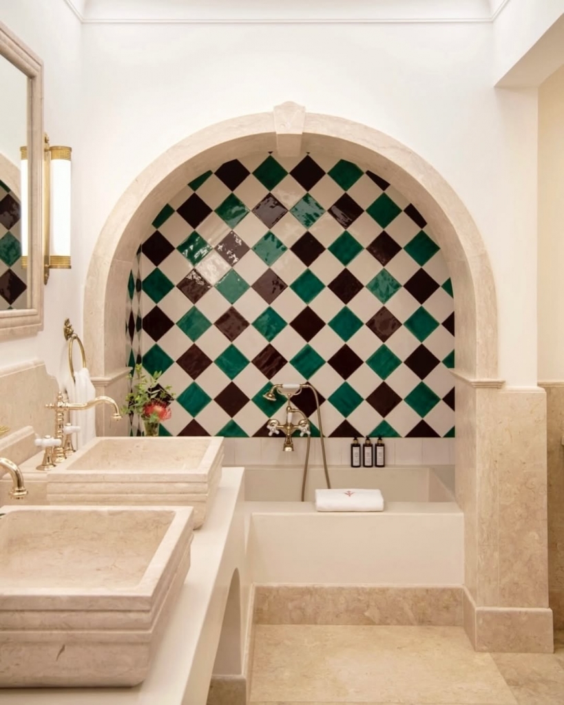 An elegant bathroom with a stone arch framing a bathtub surrounded by a dark green and white diamond-patterned tile wall, next to two stone vessel sinks. Vermelho Hotel