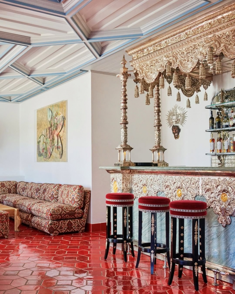 A lavish bar interior featuring a highly detailed, metallic ornate bar counter with red velvet stools, a patterned sofa, and shiny red hexagonal floor tiles. Vermelho Hotel