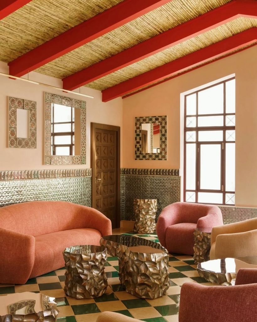 A stylish lounge with terracotta-colored curved sofas, textured metallic coffee tables, green and cream checkered floor tiles, and a bamboo ceiling with red beams. Vermelho Hotel