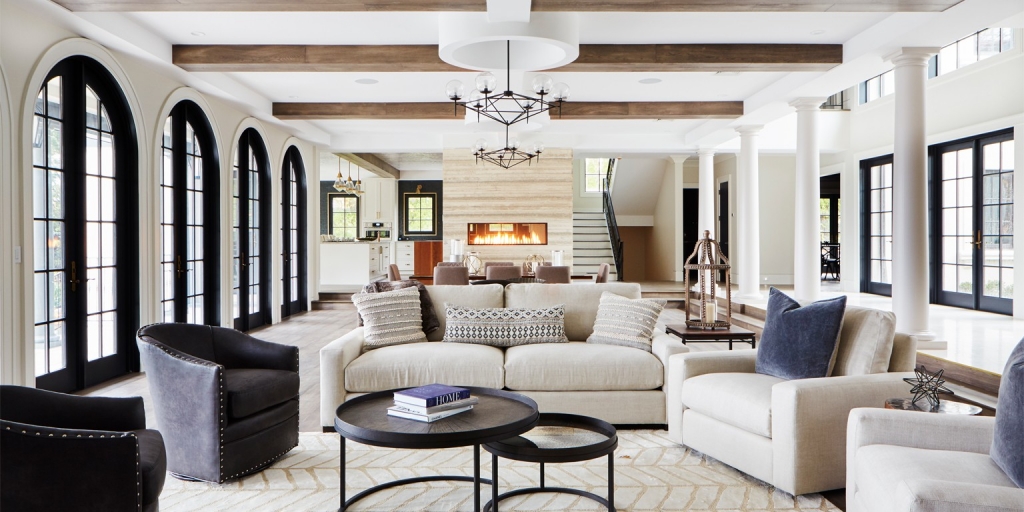 Spacious residential living room with exposed dark wood beams on the ceiling and large glass arches on the sides, focusing on a set of light-toned sofas and armchairs around black coffee tables, with a linear built-in fireplace in the background, interior designers.