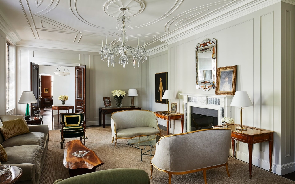 An elegant high-ceilinged room with light grey walls, a crystal chandelier, traditional white fireplace, and a mix of classic upholstered sofas and chairs, selected by interior designers