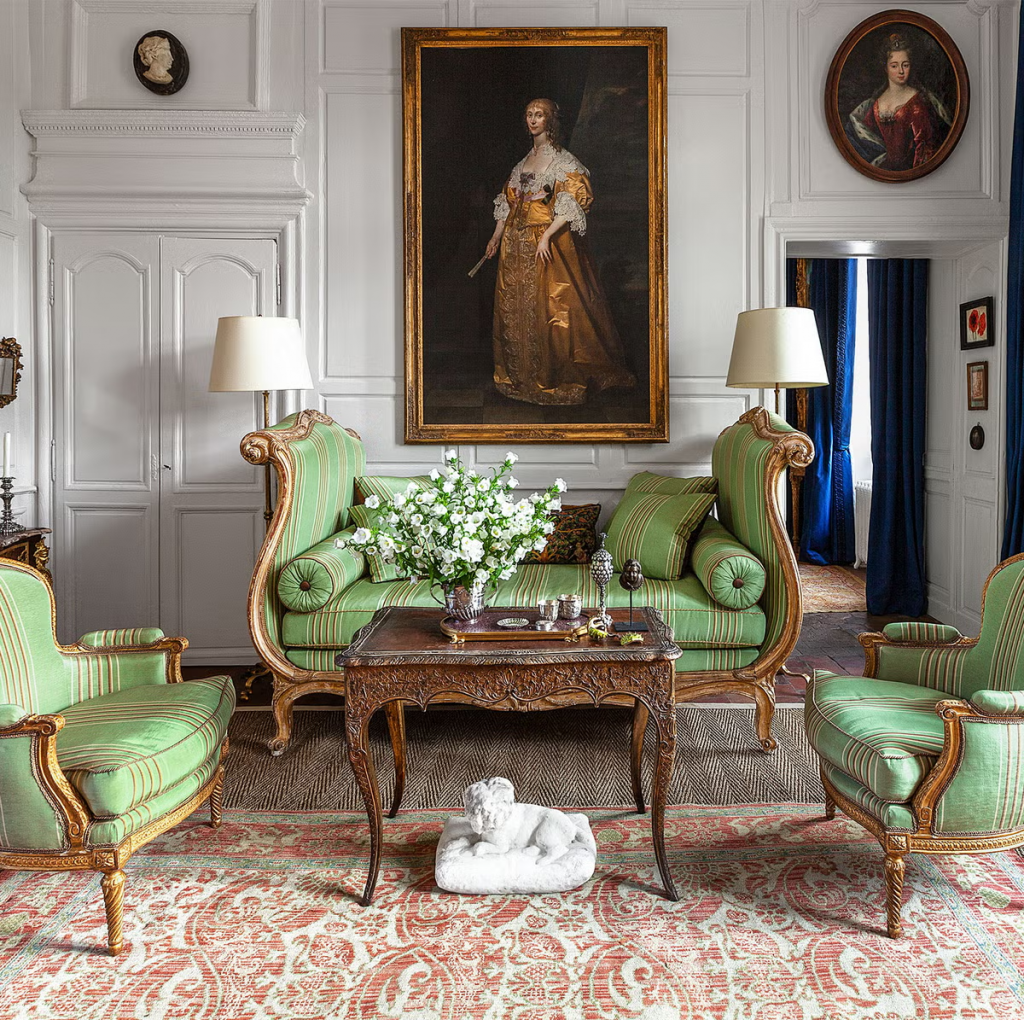 A classic, ornate European living room with white paneled walls, featuring a green and gold striped sofa and matching armchairs, a large historical portrait painting in a gold frame, and a detailed patterned rug interior designers