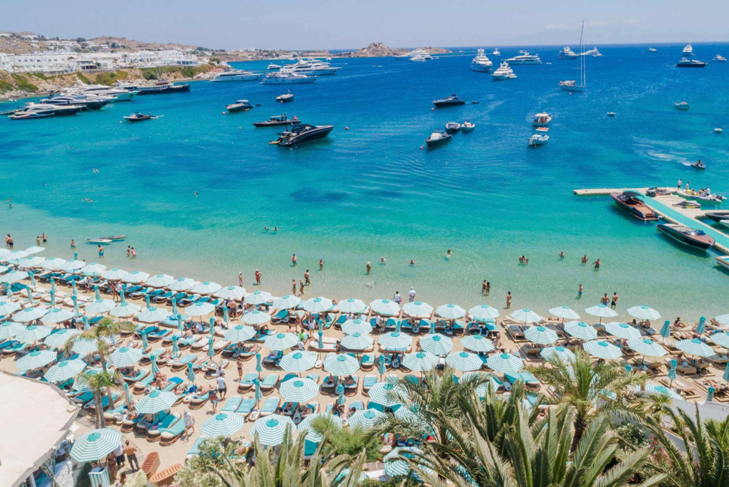 Luxury Beach Clubs: Exterior view of Nammos Mykonos on Psarou Beach, Mykonos, Greece, featuring luxurious beachfront sun loungers, elegant outdoor dining, and crystal-clear Aegean Sea views.