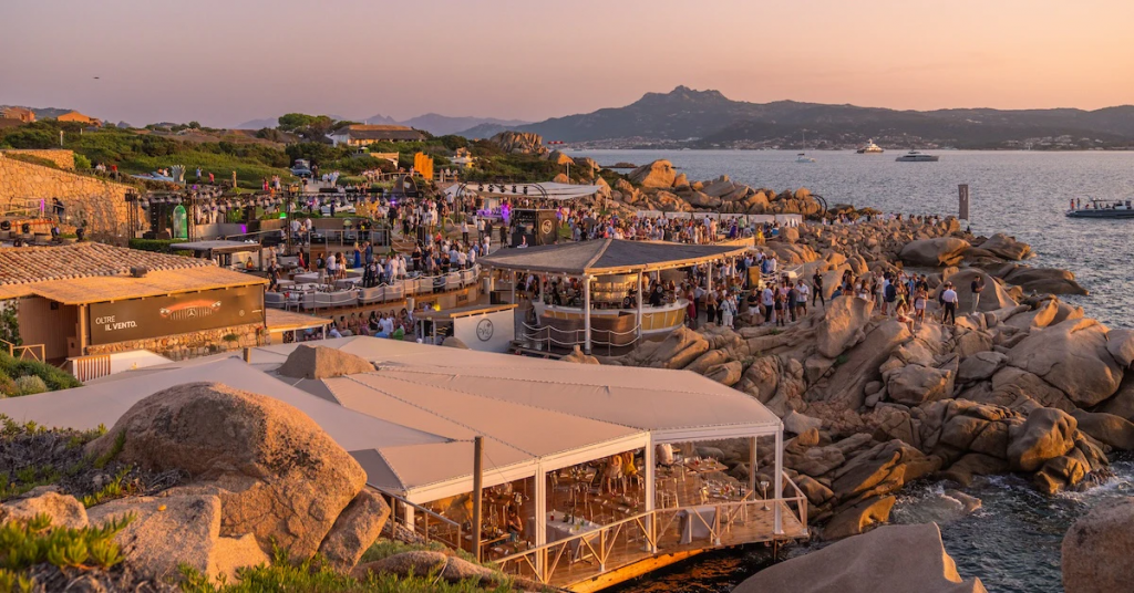 Luxury Beach Clubs: Exterior view of Phi Beach in Baja Sardinia, Costa Smeralda, Italy, featuring natural granite rock formations, elegant seaside terraces, and stunning Mediterranean sunset views.