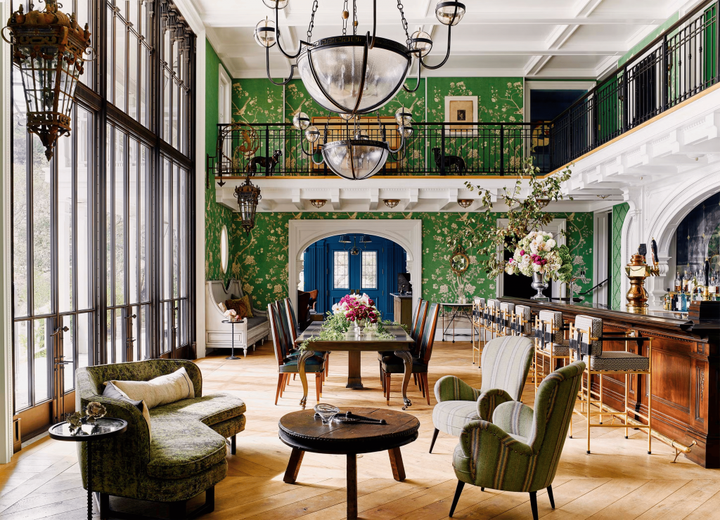 A grand, luxurious double-height dining room and bar area with green floral wallpaper, a long wooden dining table, a prominent wooden bar, elegant chandeliers, and a mezzanine balcony with a black railing interior designers