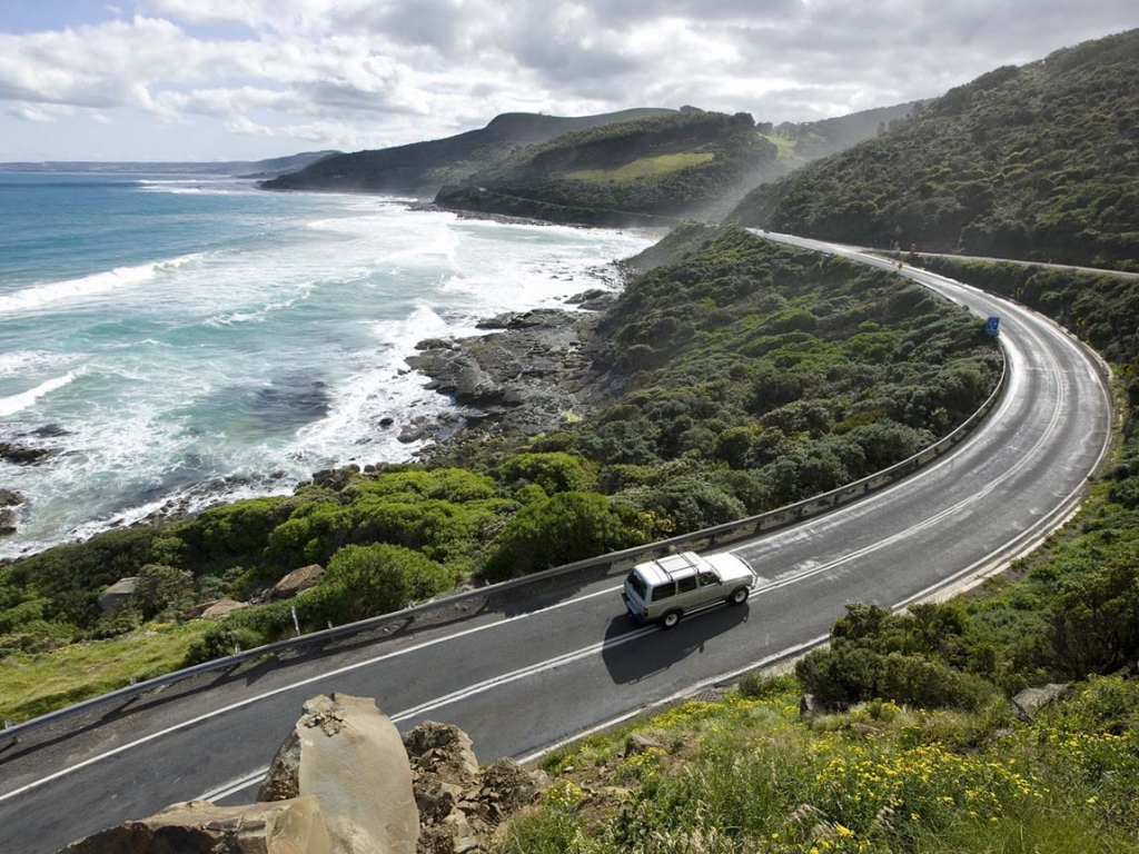 A white vehicle driving along a curved seaside highway next to a rocky, wave-swept shoreline, Coastal Roads.