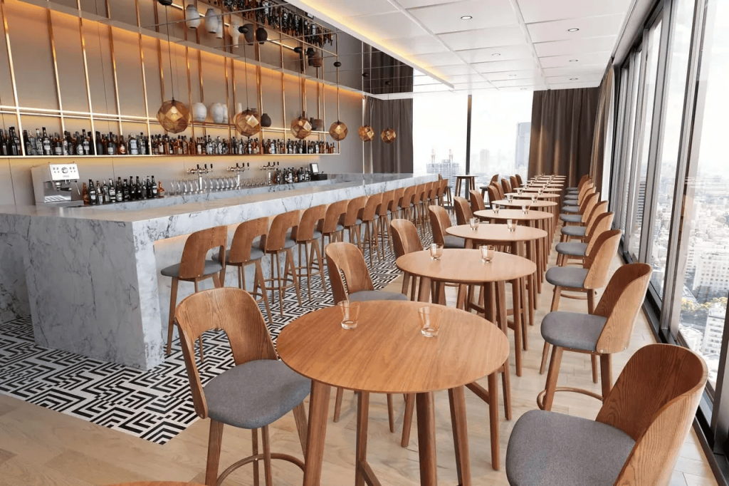 Perspective view of a luxurious hotel bar featuring a long white marble counter on the left lined with wooden barstools, and multiple round tables with upholstered chairs arranged along large panoramic windows overlooking the city, interior designers.