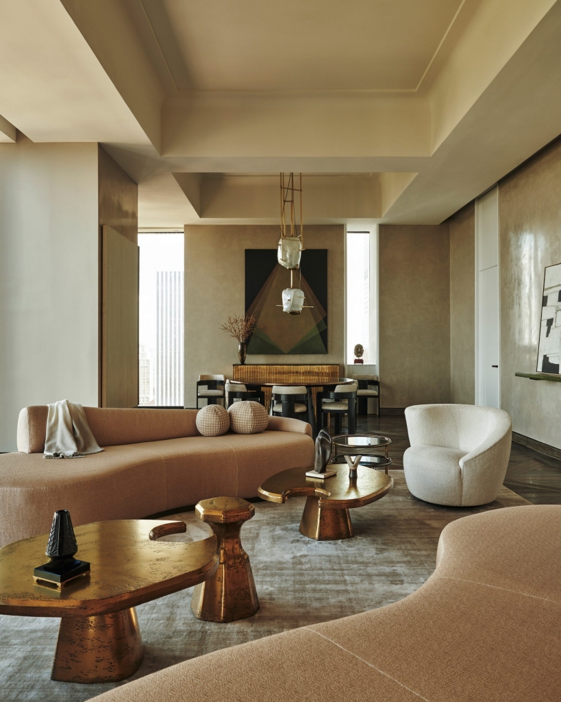 A sophisticated contemporary living room with high ceilings, featuring curved beige sofas, abstract gold coffee tables, a fluffy white armchair, and muted earth-toned walls interior designers
