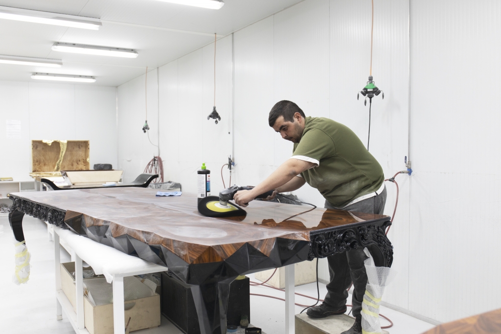 A craftsman using a power polisher on a glossy, highly detailed wooden tabletop in a workshop. Luxury Wood Furniture