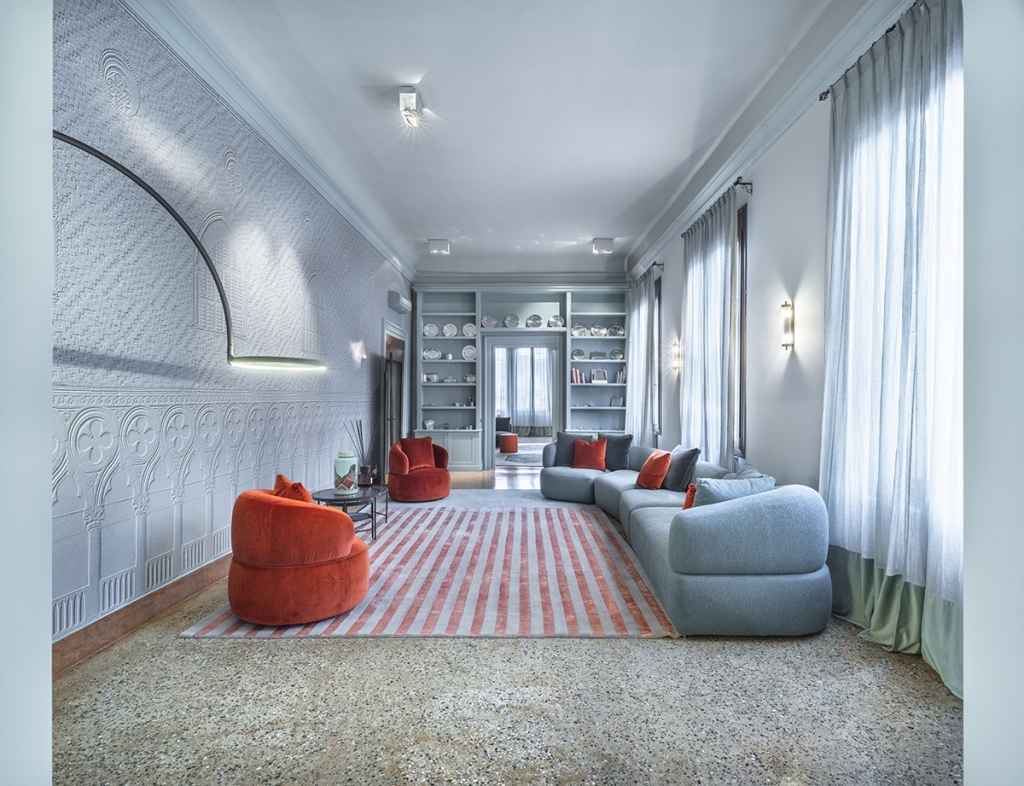 Elegant Venetian salon with textured white stucco-relief walls, a curved light-blue modular sofa, orange velvet armchairs, and a striped runner rug on terrazzo flooring. interior designers
