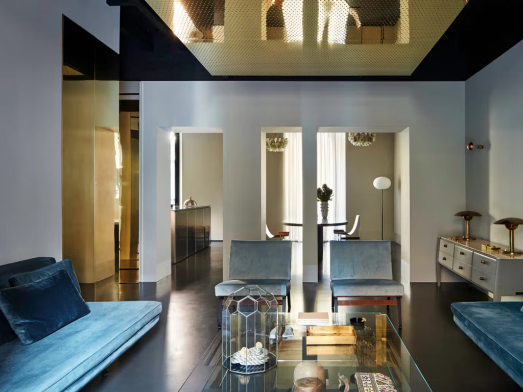 Sleek black-and-gold apartment interior with perforated brass ceiling panel, petrol blue velvet sofas, a faceted glass coffee table, and a glimpse into a dining nook beyond. interior designers