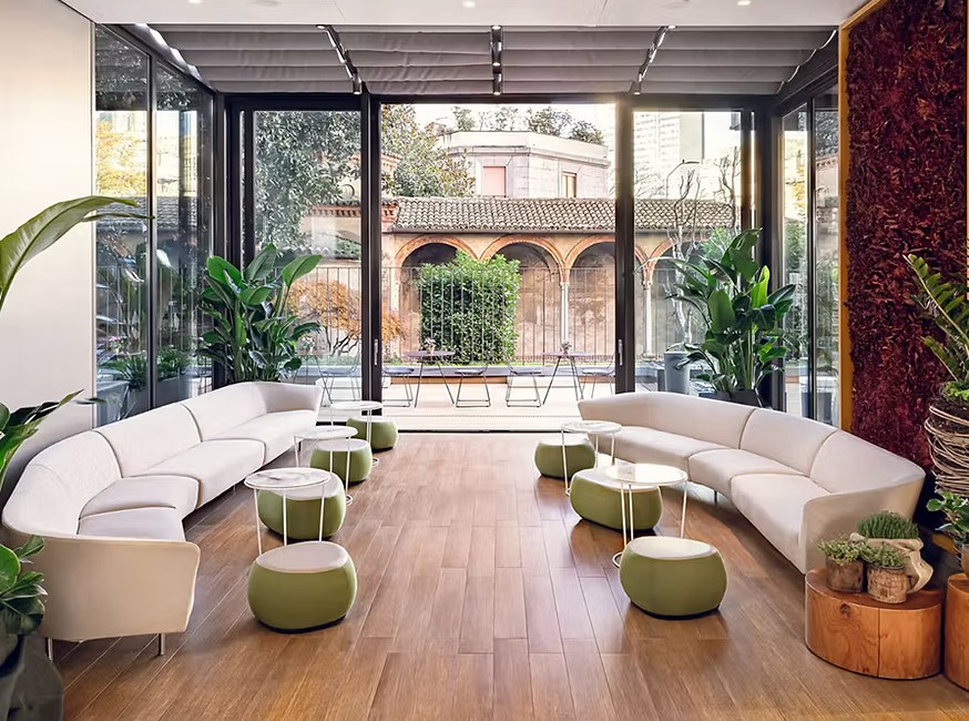 Bright glass-enclosed lounge with curved white sofas, round green ottomans, potted plants, and folding glass doors opening onto a courtyard with a historic arched cloister. interior designers