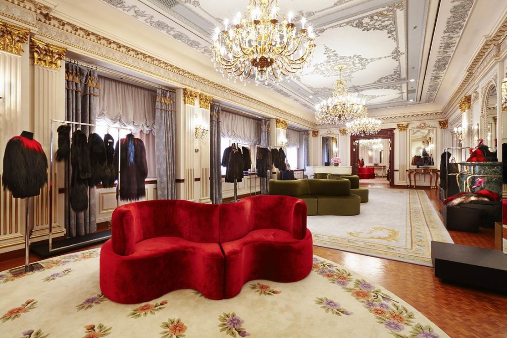 Opulent gilded showroom with ornate ceiling molding, crystal chandeliers, fur coats on display, and a curvaceous red velvet sofa on a floral rug. interior designers