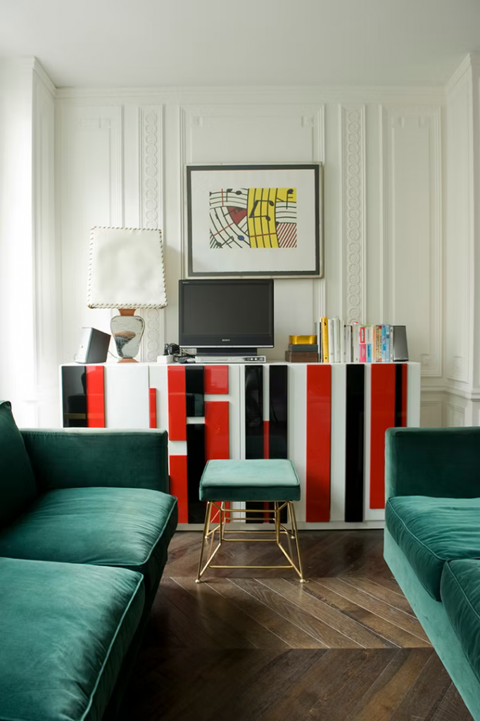Classic Parisian living room with ornate wall molding, emerald green velvet sofas, a red-and-black striped lacquered credenza, and pop-art musical print above. INTERIOR DESIGNERS