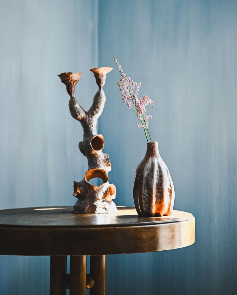 A close-up of a wooden table holding a textured bronze candelabra and a metallic ceramic vase with dried purple flowers at PAD Saint-Tropez 2026.
