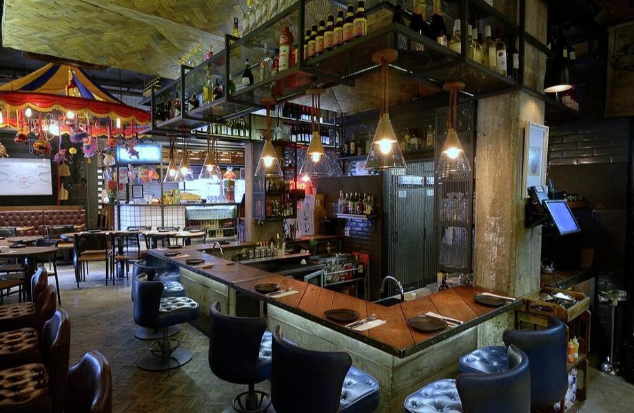 Bar decorado com prateleiras de garrafas, luminárias pendentes em formato de funil e balcão de madeira com bancos estofados em azul - melhores restaurantes em São Paulo