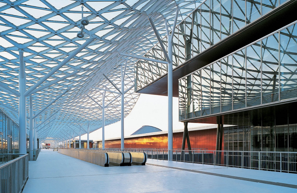 Sweeping architectural walkway with a diamond-grid glass and steel canopy roof, moving walkway below, and a glass curtain-wall building glowing amber at sunset. INTERIOR DESIGNERS